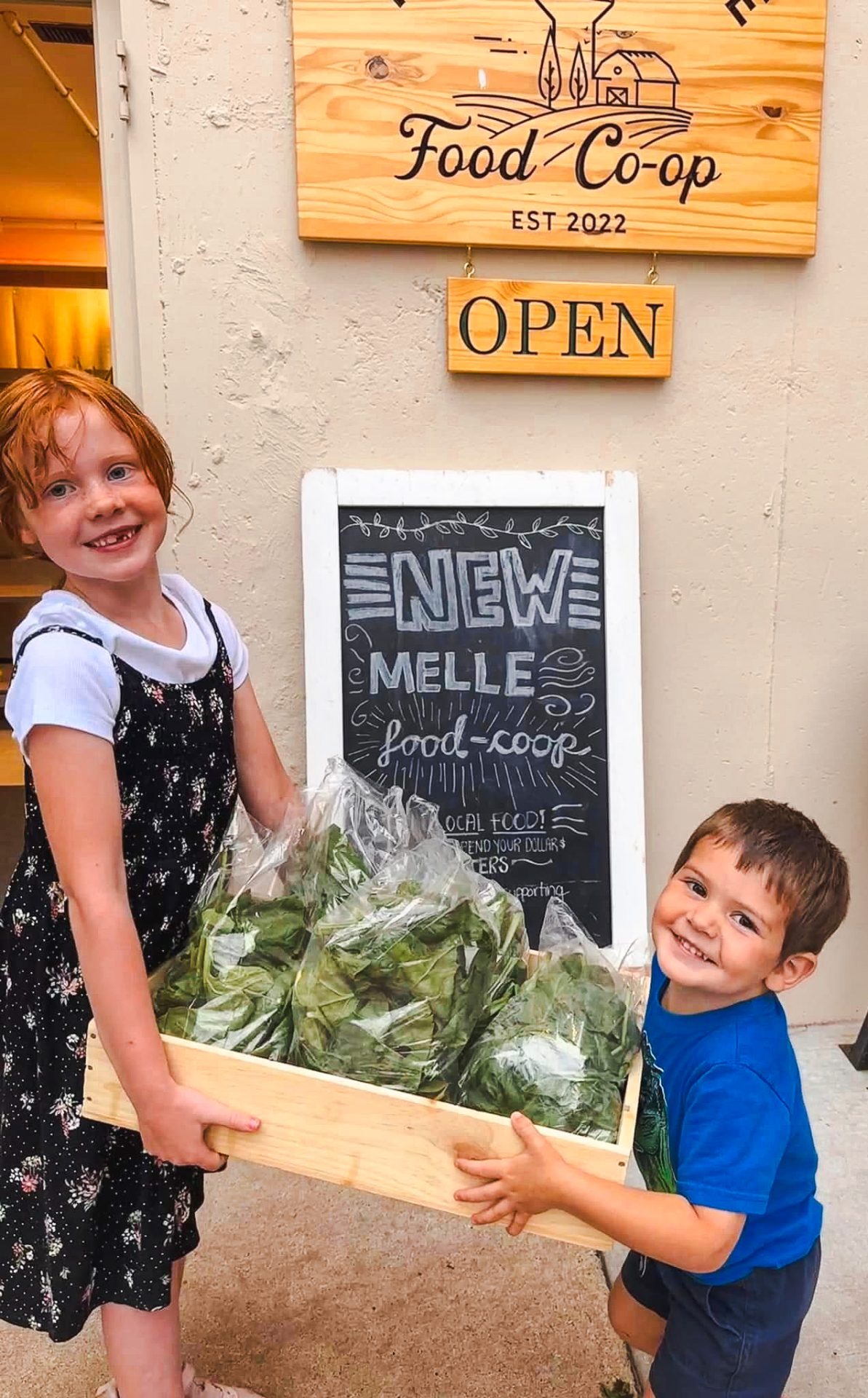Two young children outside the New Melle Food Co-op entrance holding a box with bags of spinach